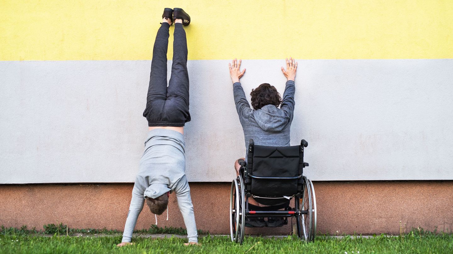Zwei Personen mit dem Rücken zum Betrachter vor einer zweifarbigen Wand, auf einer Grasfläche. Die Person rechts sitzt im Rollstuhl und stützt die Arme an der Wand ab, die Person links macht einen Handstand an der Wand. Zwei Personen mit dem Rücken zum Betrachter vor einer zweifarbigen Wand, auf einer Grasfläche. Die Person rechts sitzt im Rollstuhl und stützt die Arme an der Wand ab, die Person links macht einen Handstand an der Wand.