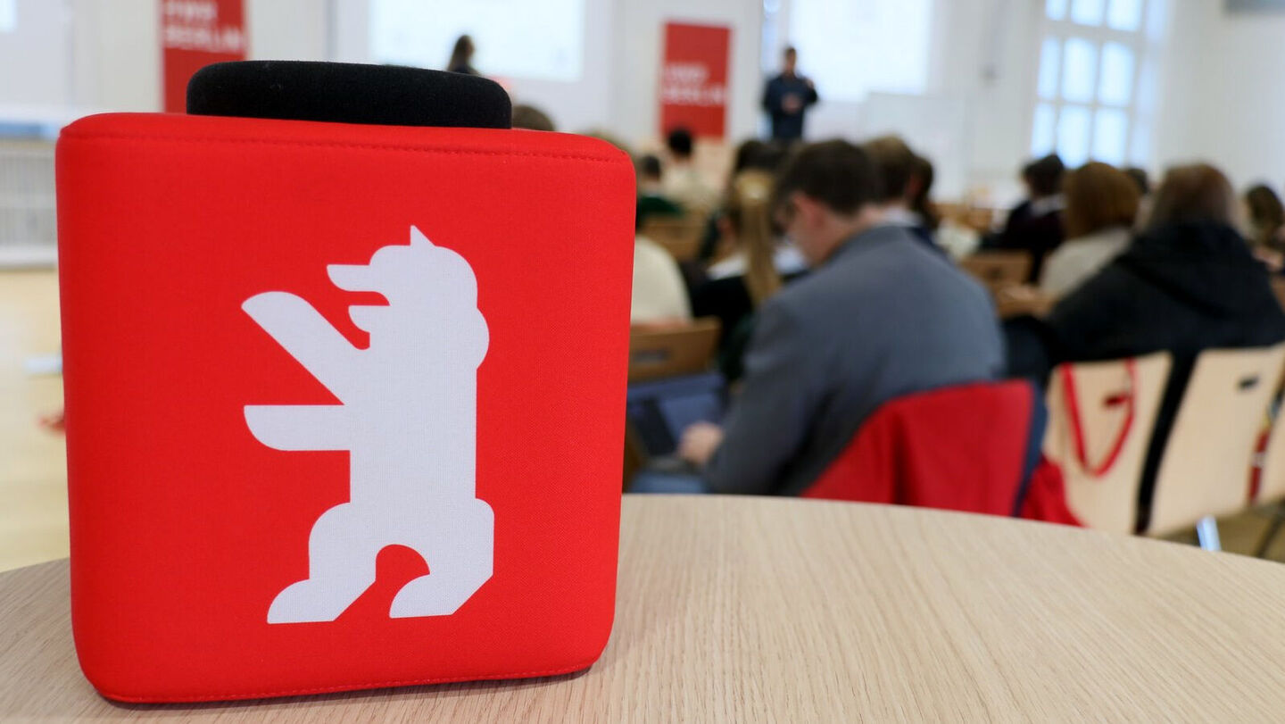 Rotes Quadrat mit weißem Berliner Bären auf einem Tisch vor unscharfem Publikum in einem hellen Raum.