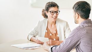 [Translate to English:] Student Counseling: Counselor with curly hair and glasses at the table talking to a student in a checkered shirt. Photo: © kate_sept2004/E+/Getty Images