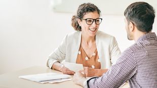 Studienberatung: Beraterin mit lockigem Haar und Brille am Tisch im Gespräch mit einem Studenten mit kariertem Hemd. Foto: © kate_sept2004/E+/Getty Images