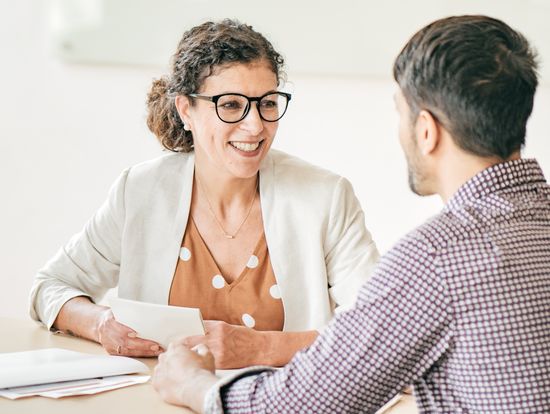 Studienberatung: Beraterin mit lockigem Haar und Brille am Tisch im Gespräch mit einem Studenten mit kariertem Hemd. Foto: © kate_sept2004/E+/Getty Images