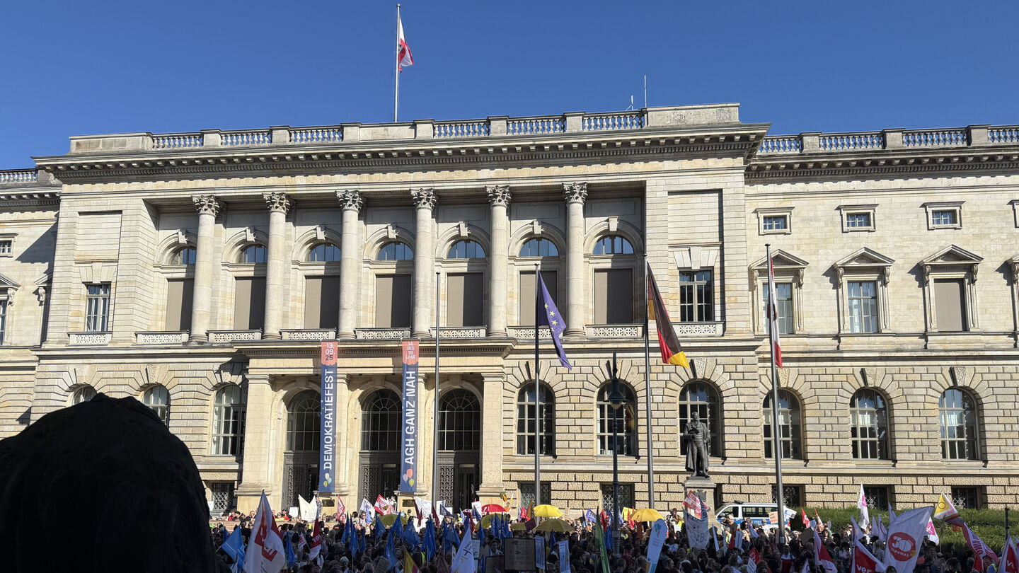Menschenmenge mit Protestschildern vor einem historischen Gebäude mit Säulen und Flaggen