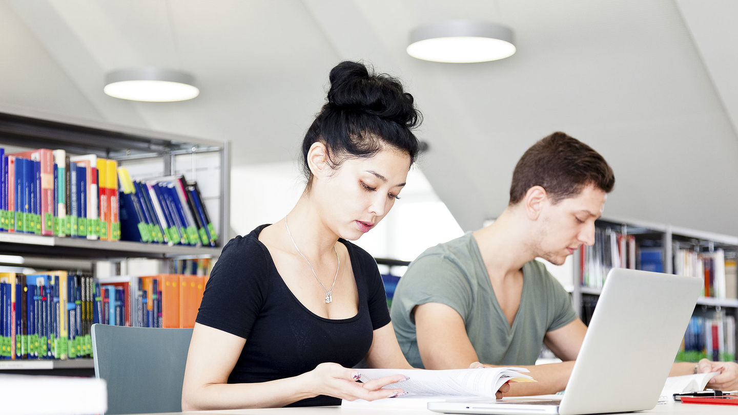 Studierendenportal: Eine Studierende und ein Studierender arbeiten vor ihren Laptops in der Hochschulbibliothek der HWR Berlin. Im Hintergrund Regale. Foto:  © Klaus Lange photography