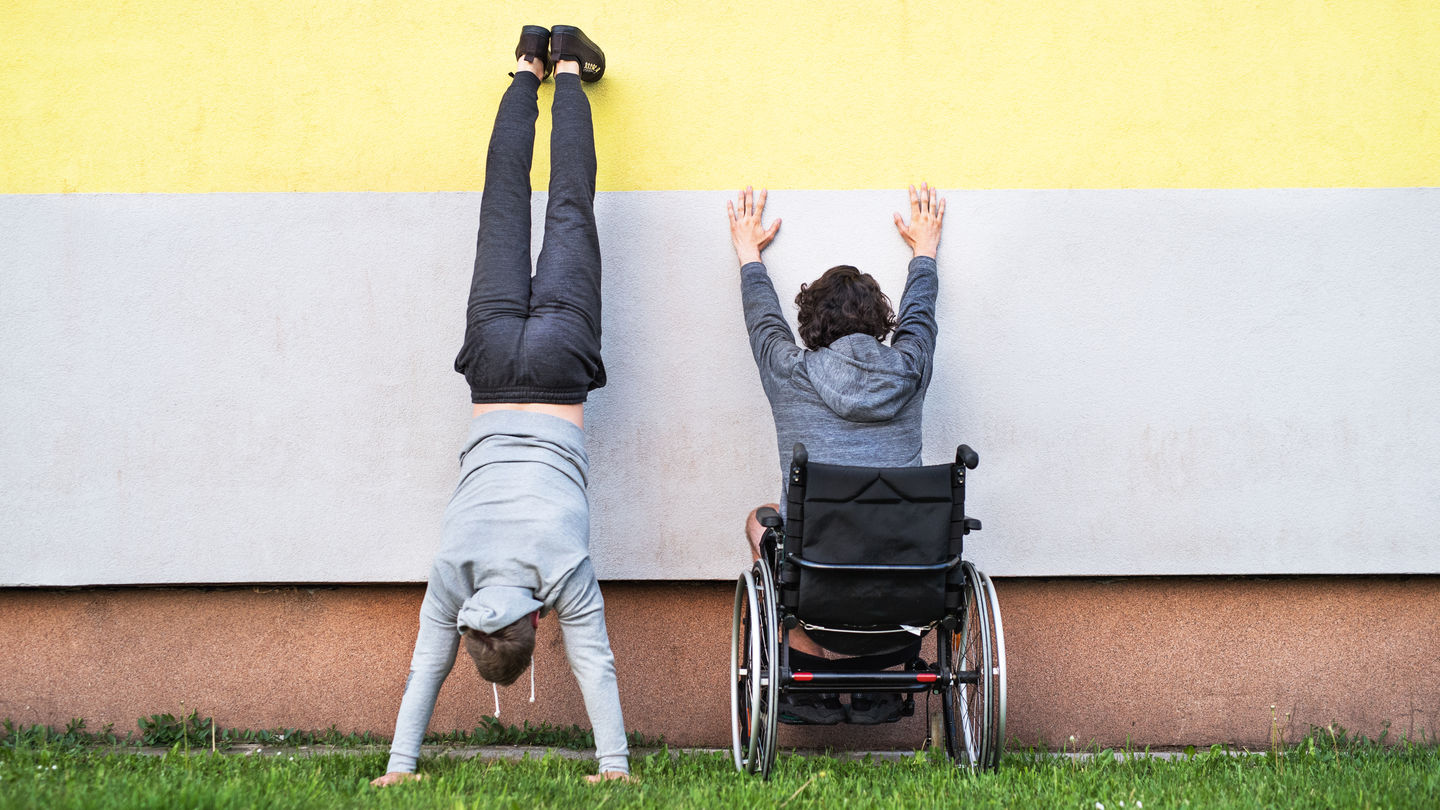 Zwei Personen mit dem Rücken zum Betrachter vor einer zweifarbigen Wand, auf einer Grasfläche. Die Person rechts sitzt im Rollstuhl und stützt die Arme an der Wand ab, die Person links macht einen Handstand an der Wand.