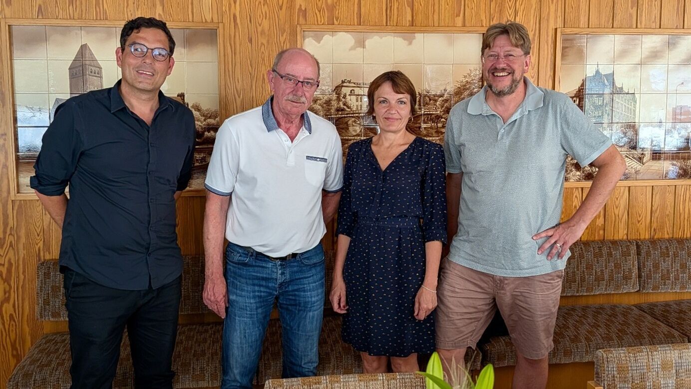 Vier Personen stehen nebeneinander vor einer Wand mit Holzvertäfelung und einem Bild mit Stadtansicht, zwei Männer tragen Brillen, eine Frau trägt ein dunkelblaues Kleid mit Punkten.