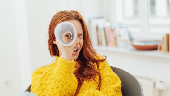 Junge Frau in gelben Pullover schaut durch ein zusammengerolltes Papier wie durch ein Fernrohr.