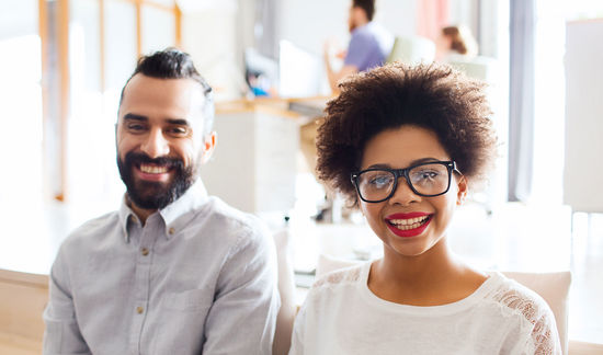 A young woman with glasses and a young man with dark hair and beard, who are not from Germany, sit on the sofa and laugh into the camera. Photo: Getty Images