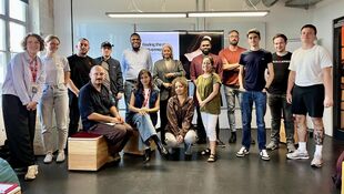 Group of people in a room in front of a screen, some sitting on stools and others standing, all looking at the camera
