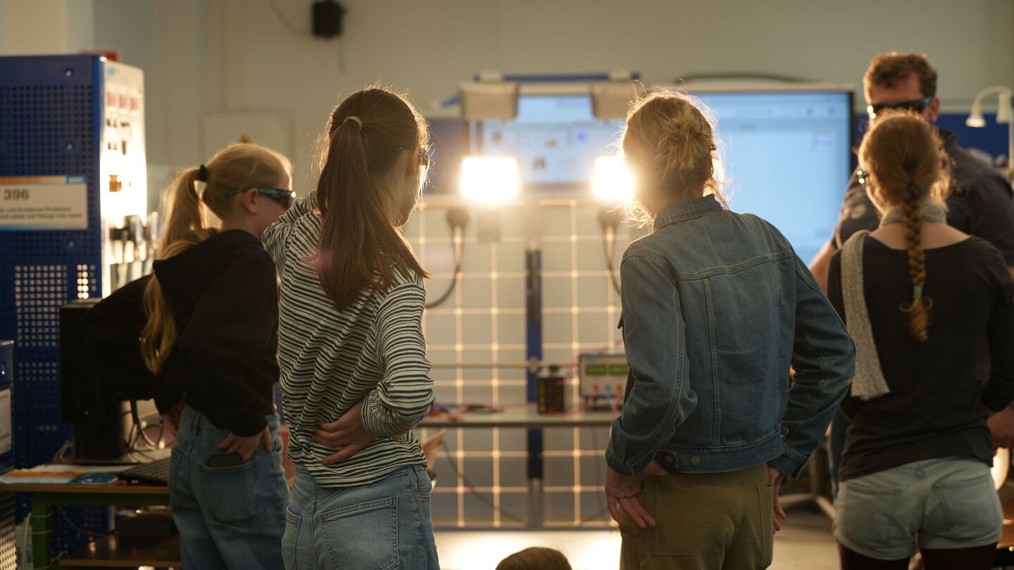 Gruppe von Mädchen steht mit dem Rücken zur Kamera und blickt auf eine helle Lichtquelle in einem Innenraum.