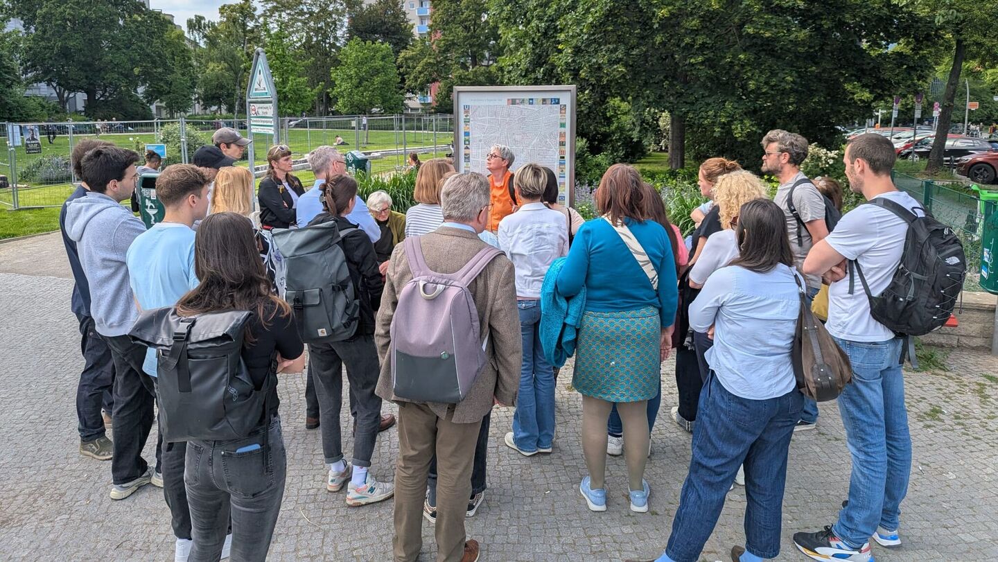 Gruppe von Menschen steht im Freien vor einer Informationstafel auf einem gepflasterten Weg, umgeben von Bäumen und Gebäuden