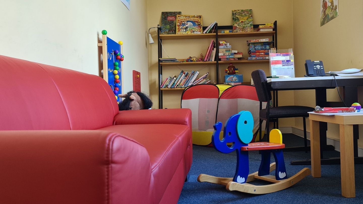 Kinderspielzimmer mit rotem Sofa, blauem Schaukelelefant, Bücherregal und Tischen mit Stühlen.