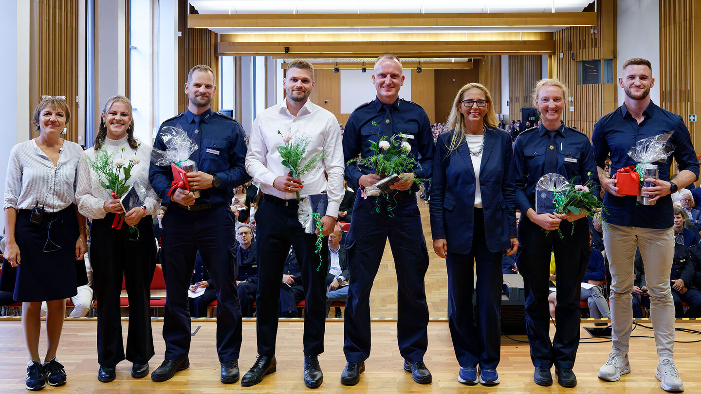 Sieben Personen stehen nebeneinander in einem Saal, einige tragen Uniformen, andere halten Blumen und Geschenke in den Händen.