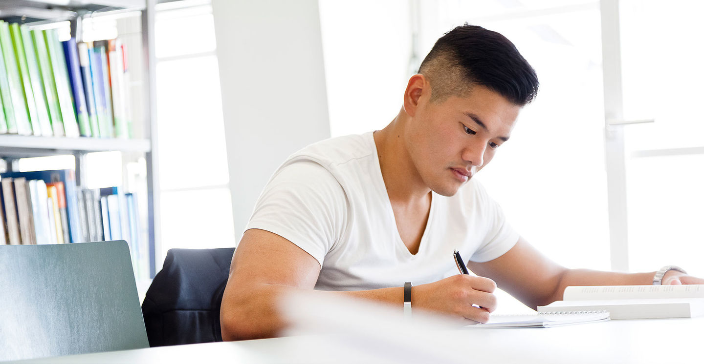 International studieren: Ein internationaler Student aus dem asiatischen Raum sitzt in der Bibliothek an einem Arbeitstisch und macht sich Notizen. Foto: © Klaus Lange photography