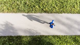 Vogelperspektive: Ein Studierender in blauer Sportkleidung joggt auf einem Asphaltweg über eine grüne Wiese. Foto: © GrapeImages/Getty Images/iStockphoto
