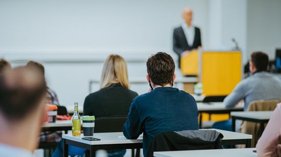 Lehrsaal: Ein Mann steht vorne am Pult, vor im sitzen Studierende. 