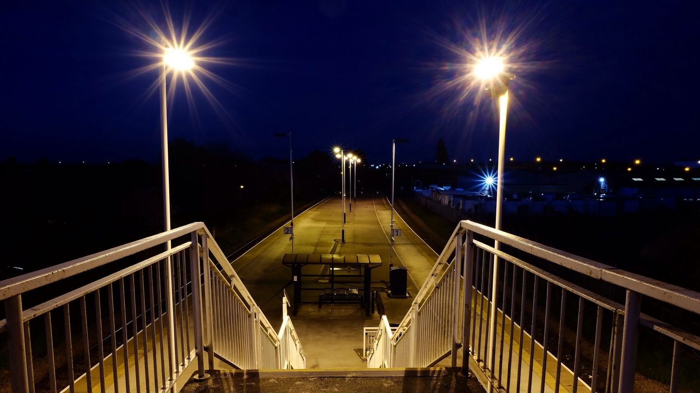 Blick von einer beleuchteten Treppe auf einen nächtlichen, von Straßenlaternen gesäumten Bahnsteig mit einer Bank in der Mitte