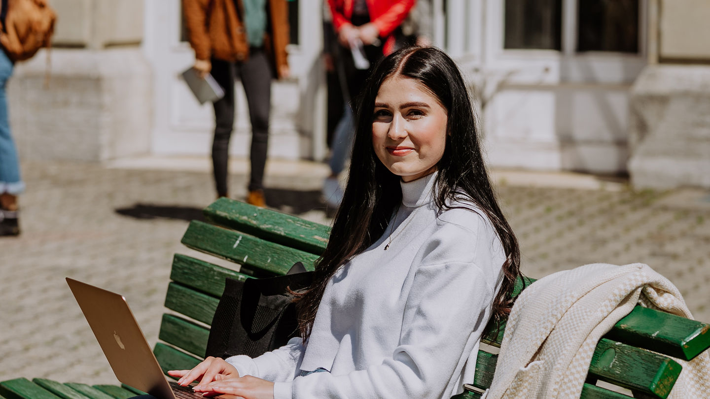 Studienbeginn an der HWR Berlin: Infos für Ersties zum Studienstart. Foto: Lukas Schramm Eine Studierende der HWR Berlin sitzt mit dem Laptop auf dem Schoß auf einer grünen Bank und lächelt in die Kamera. Foto: Lukas Schramm