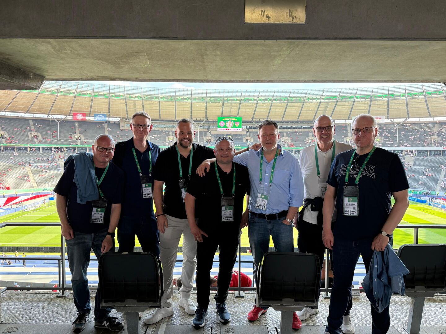 Sieben Männer stehen in einer Stadionloge mit Blick auf ein Fußballfeld, alle tragen Ausweise um den Hals
