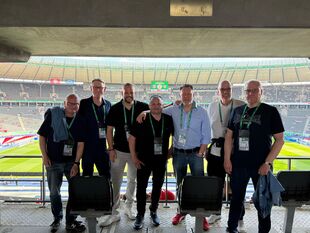 Sieben Männer stehen in einer Stadionloge mit Blick auf ein Fußballfeld, alle tragen Ausweise um den Hals