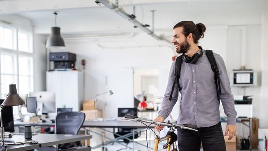 Bewerbung duales Studium: Dual Studierender mit Bart, Kopfhörer um den Hals und grauem Hemd schiebt sein Fahrrad im Büro seines Unternehmens.