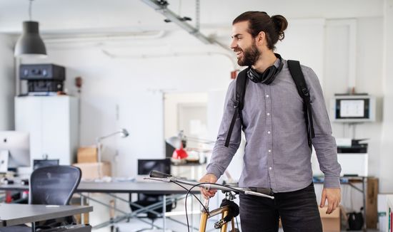 Bewerbung duales Studium: Dual Studierender mit Bart, Kopfhörer um den Hals und grauem Hemd schiebt sein Fahrrad im Büro seines Unternehmens. Foto: © alvarez/E+/Getty Images