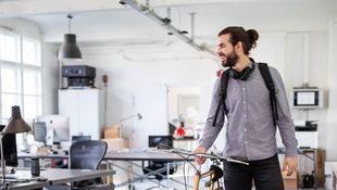 Application for cooperative studies: Cooperative student with a beard, headphones around his neck and a gray shirt pushes his bicycle in his company's office.