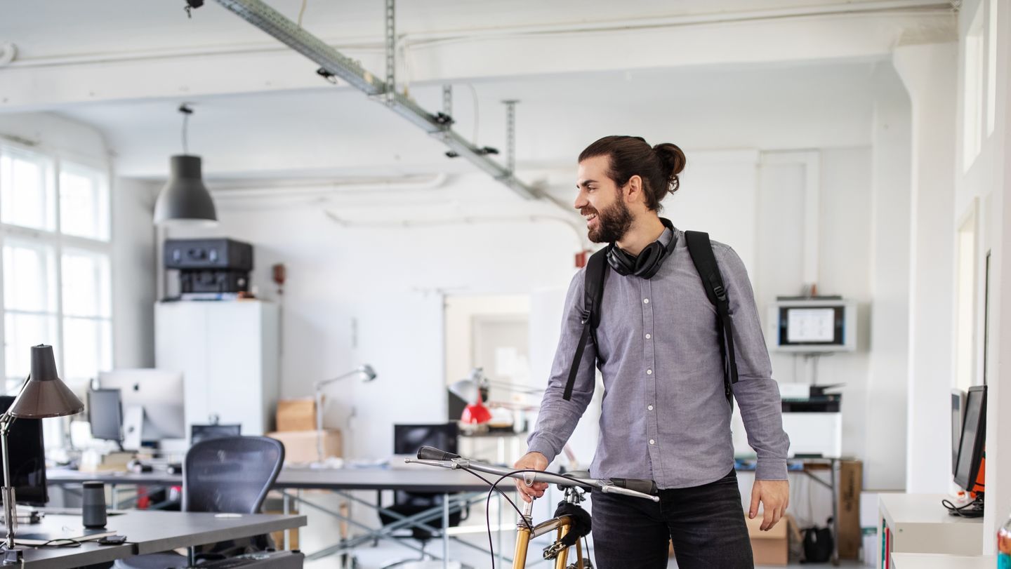 Application for cooperative studies: Student with beard, headphones around his neck and grey shirt pushing his bike in his company's office.  Photo: © alvarez/E+/Getty Images