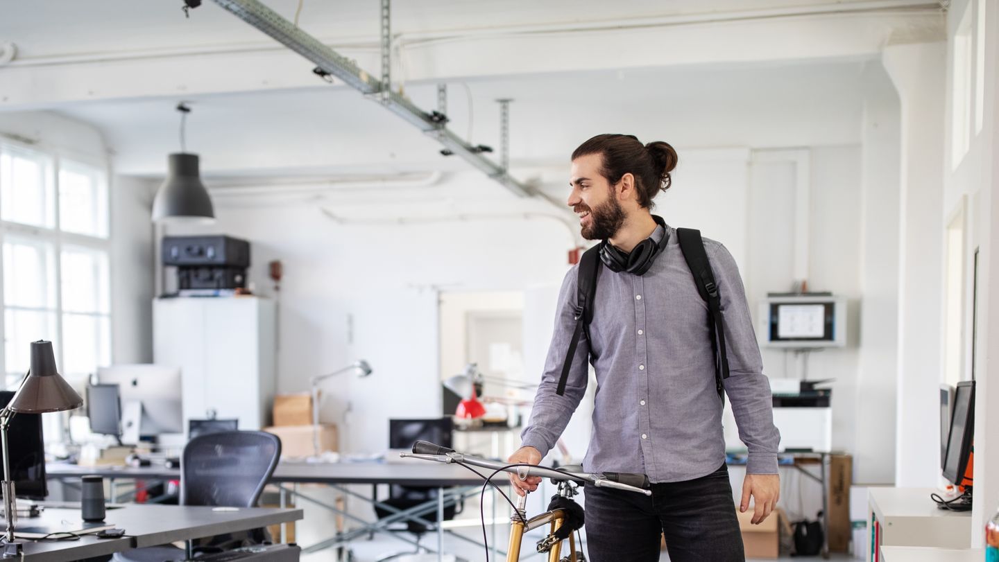 Bewerbung duales Studium: Dual Studierender mit Bart, Kopfhörer um den Hals und grauem Hemd schiebt sein Fahrrad im Büro seines Unternehmens. Foto: © alvarez/E+/Getty Images